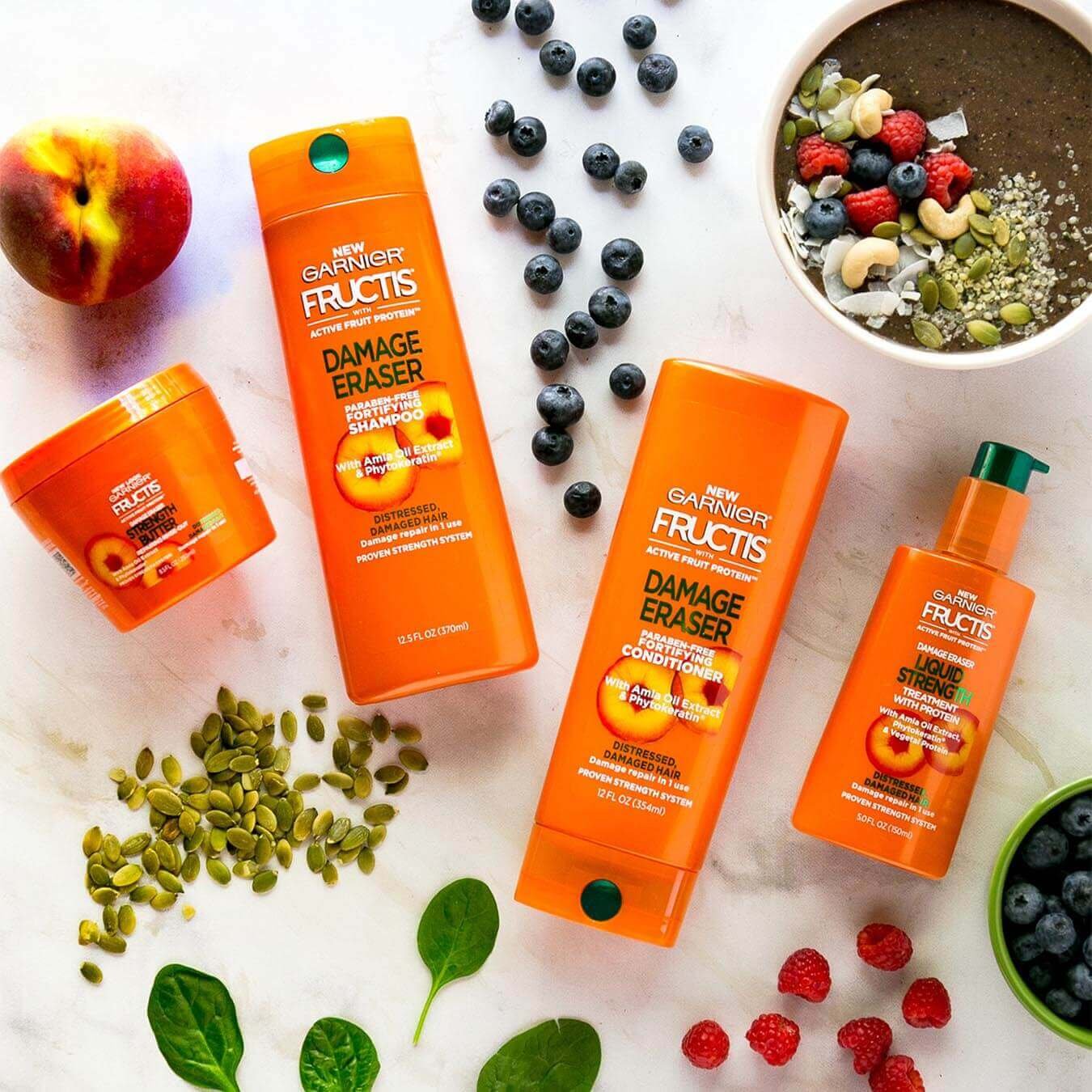 Garnier Fructis Damage Eraser Shampoo and Damage Eraser Conditioner on white marble next to raspberries, blueberries, basil leaves, pumpkin seeds, and soil with various toppings.